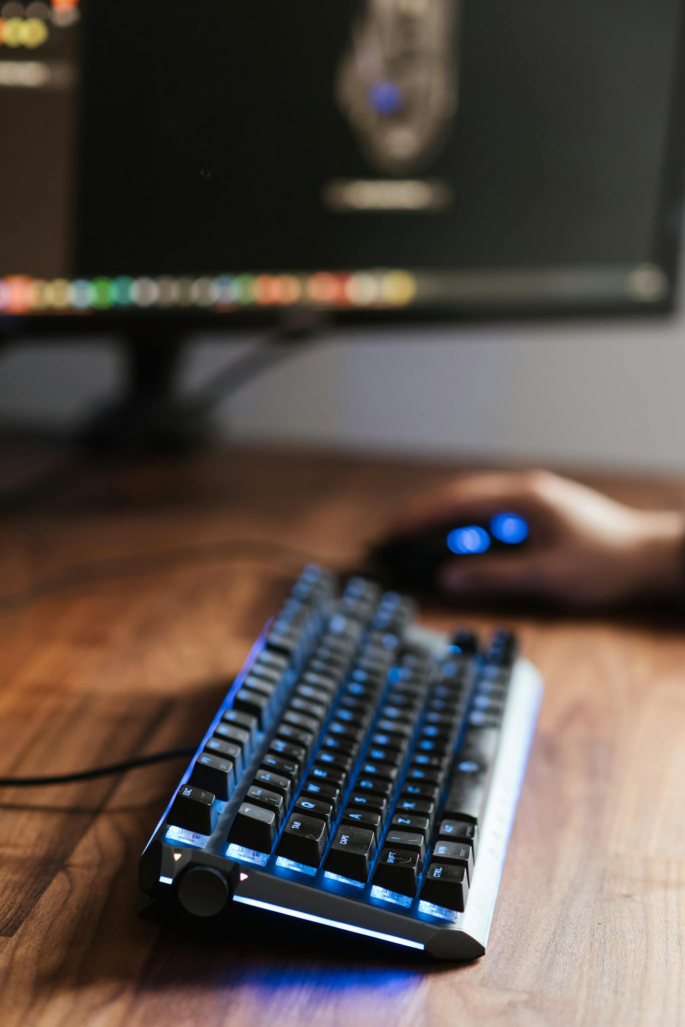 Crop unrecognizable person sitting at table with computer monitor and backlit keyboard while using mouse at home